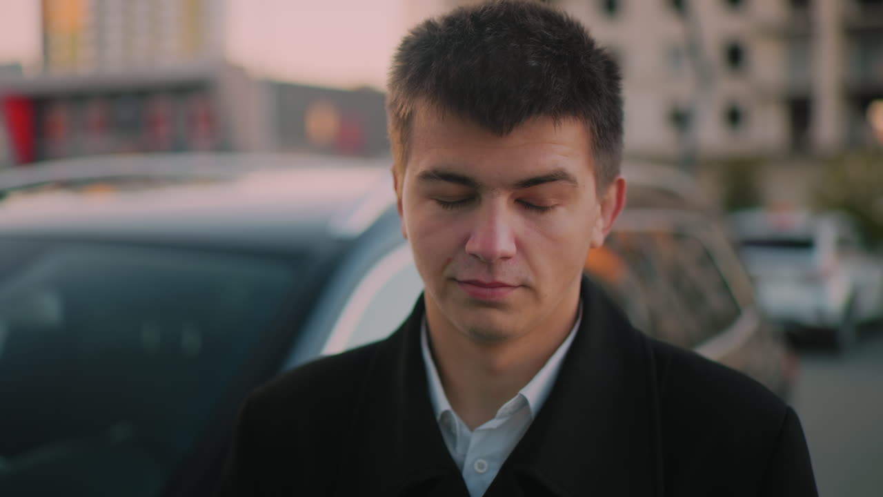 Man standing close to car with subtle smile, blurred background shows parked cars and distant building at dusk, portraying confident business professional enjoying success in urban environment