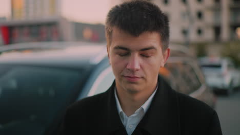 man standing close to car with subtle smile, blurred background shows parked cars and distant building at dusk, portraying confident business professional enjoying success in urban environment