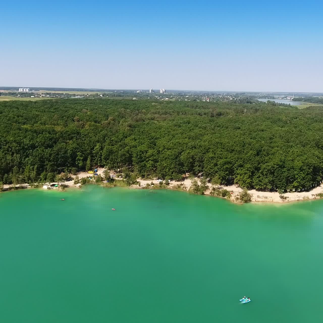 Deep blue lake with white sandy banks around. Green forest covering the waterfront. Cityscape and clear sky at the backdrop