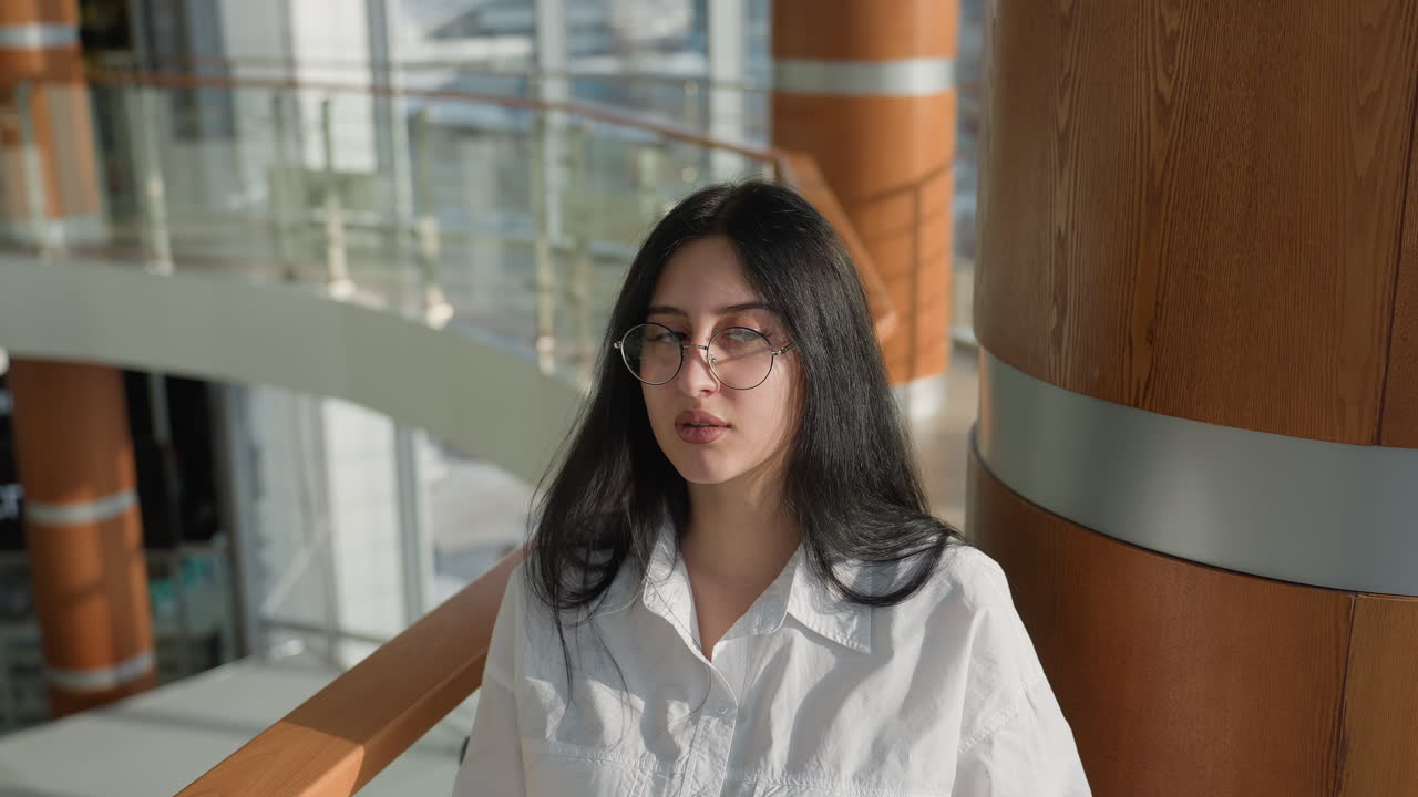 Businesswoman in white shirt and glasses standing by glass railing in spacious contemporary mall, patiently looking around while waiting for client