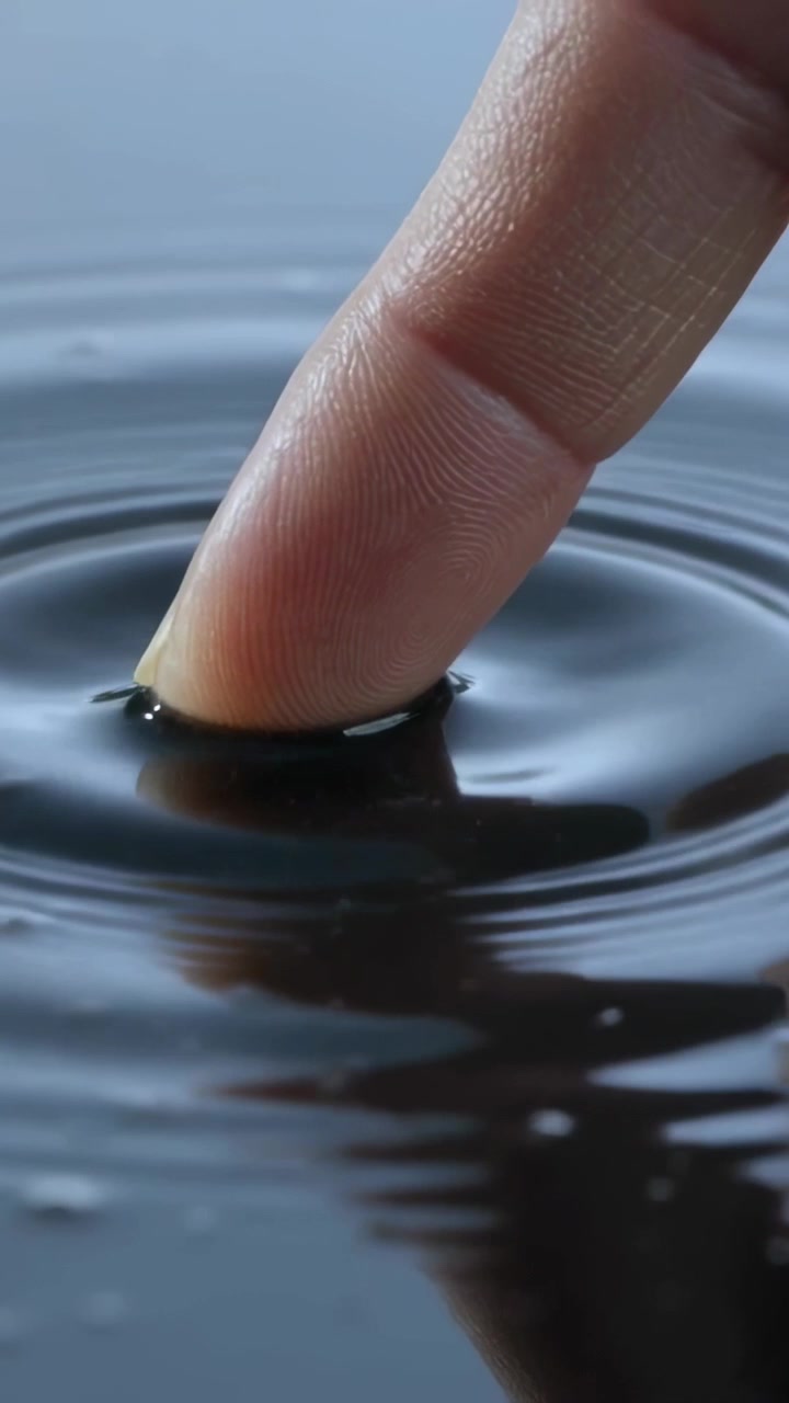Video verticale: Punta del dito che preme sull'acqua scura, creando increspature, distorcendo il riflesso