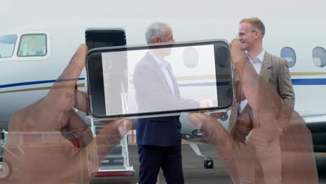 video compuesto de una mano sosteniendo un teléfono inteligente sobre dos hombres de negocios que se estrechan la mano en el aeropuerto