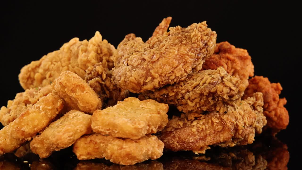 Golden fried chicken wings, drumsticks, and nuggets rotate on a reflective surface under warm studio lighting, highlighting crunchy textures and appetizing detail