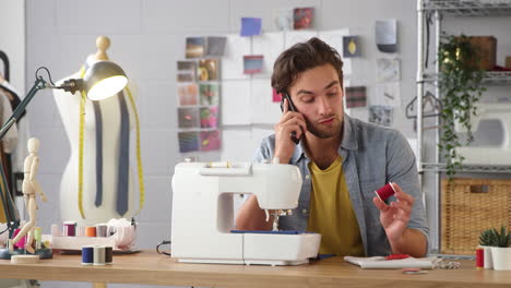 male owner of fashion business making call on mobile phone at sewing machine in studio
