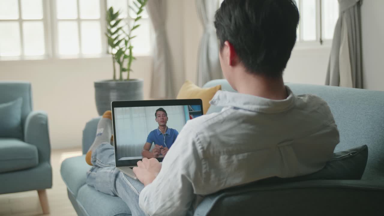 自宅のリビングルームでノートパソコンから同僚とビデオ会議で挨拶するアジア人男性、4K動画