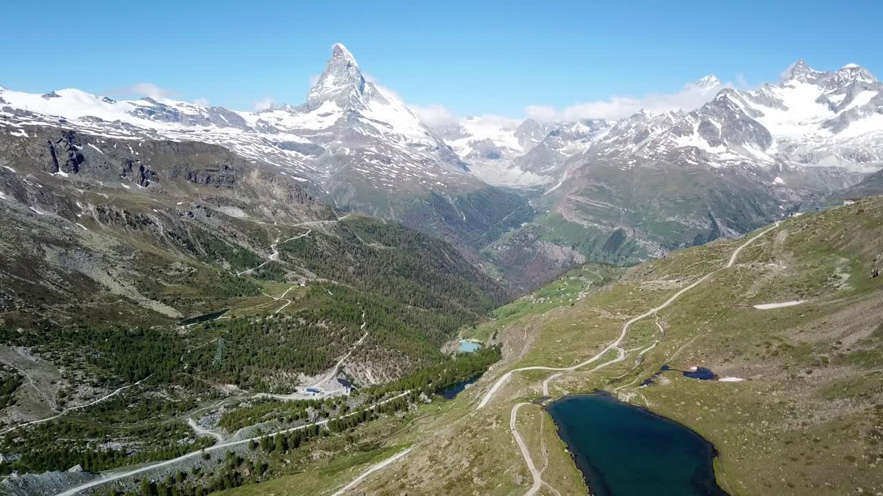 Aerial view of Zermatt, showcasing the iconic Matterhorn mountain surrounded by snow-capped peaks, lush green valleys, winding roads, and a serene alpine lake in the foreground