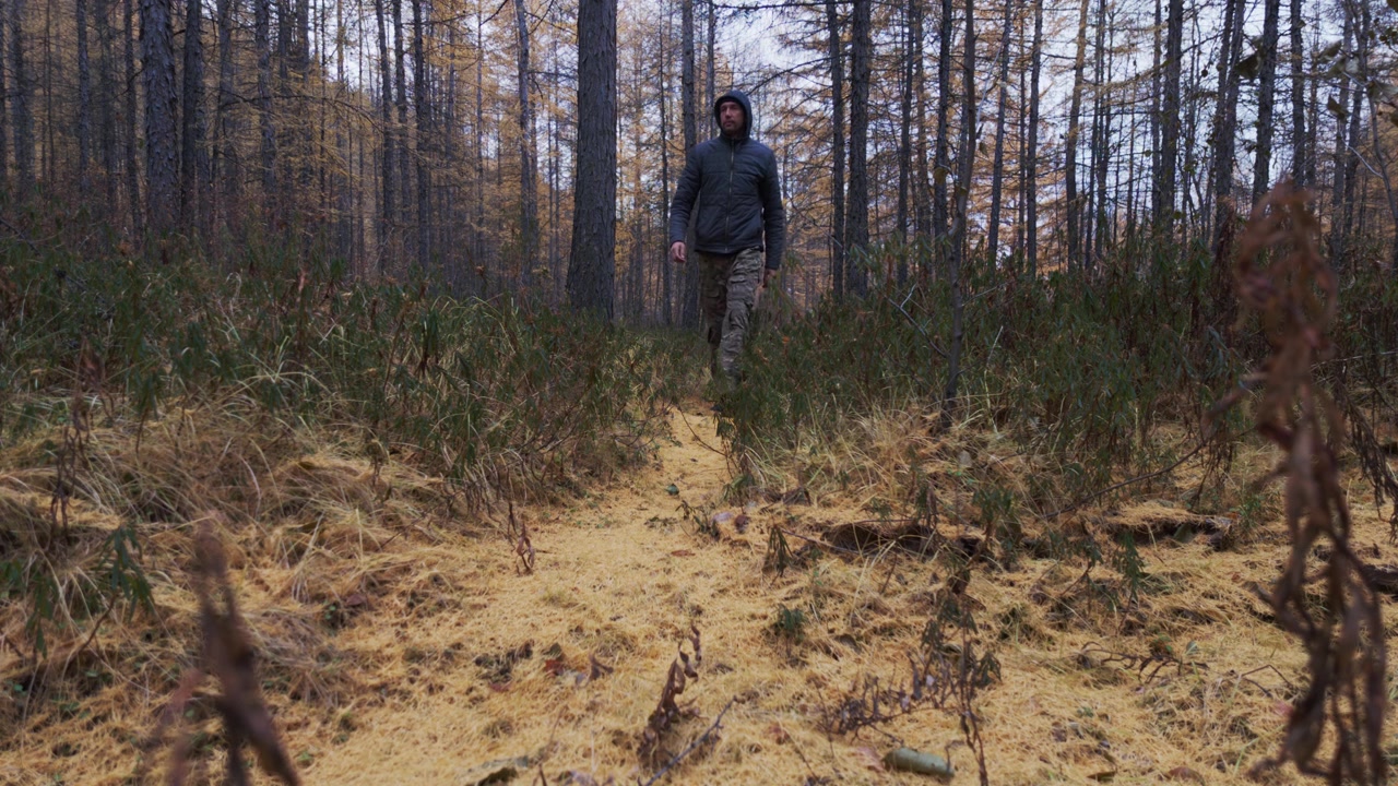 A man in a dark hooded jacket and camouflage pants walks steadily along a narrow dirt path through a forest with tall, thin trees and dry underbrush.