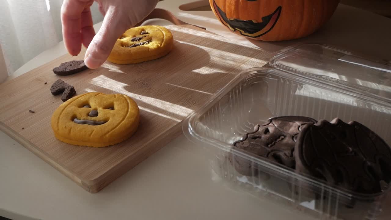 Side view of hand placing bat and pumpkin-shaped Halloween cookies on wooden board beside pumpkin under sunlight