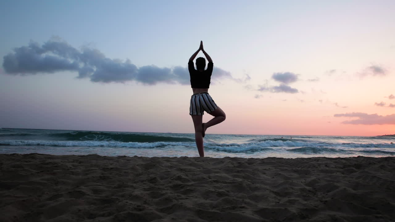 Brunette with wide hips and stripped shorts doing yoga tree pose on the beach at sunset.