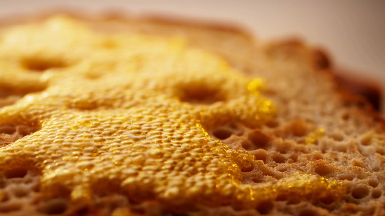 Scena ravvicinata in movimento che mostra il miele che scorre su un favo di cera d'api naturale, passando da goccioline separate a strati lucidi continui che collegano le celle, catturano la luce calda e rivelano una ricca texture