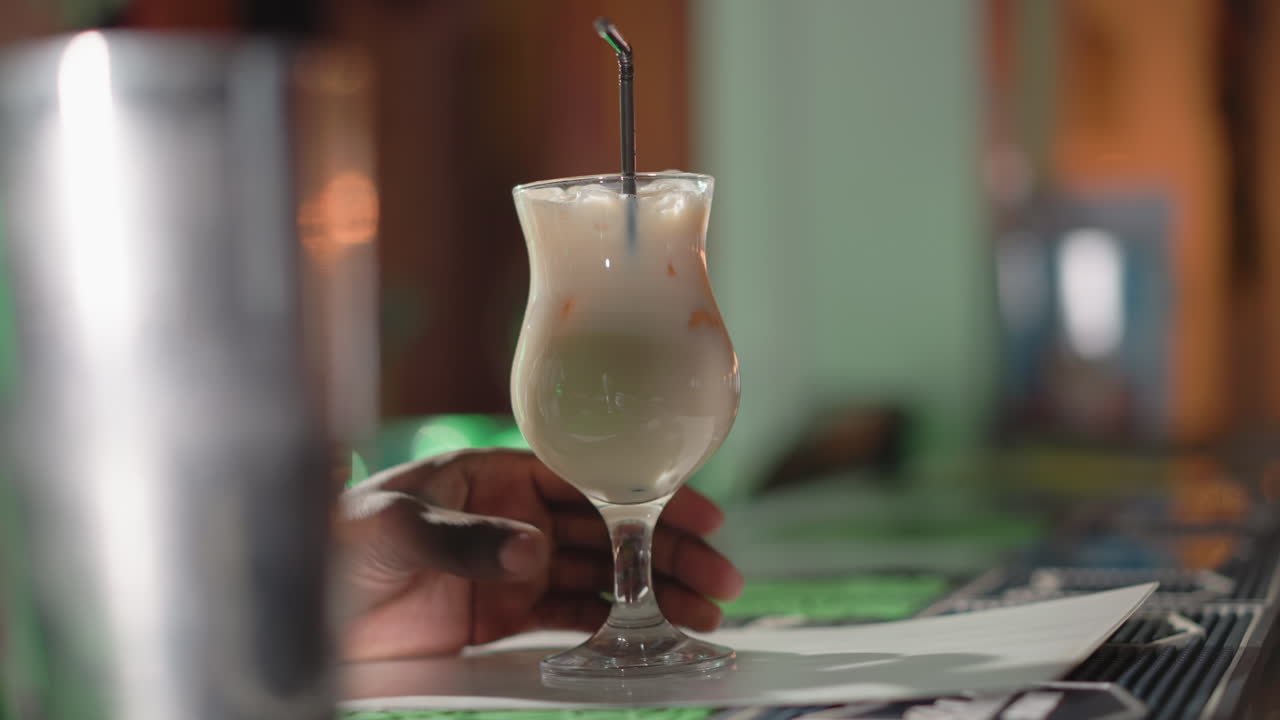Hand stirring iced drink in elegant glass with straw on bar counter. The focus is on the drink with ice cubes and a blurred background, showcasing a vibrant bar atmosphere