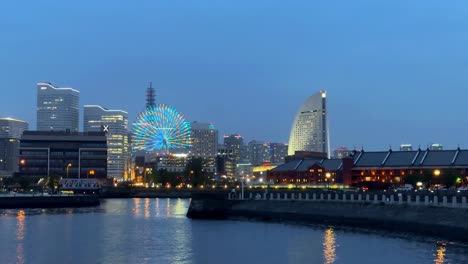 Abendliche-Skyline-Mit-Beleuchtetem-Riesenrad-Am-Wasser,-Lichter-Der-Stadt-Spiegeln-Sich-Im-Wasser,-Sonnenuntergang