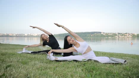 two young women are doing yoga while sitting on the shore of the lake two young women are doing yoga while sitting on the shore of the lake against the background of the sky yoga in the fresh air on the shore of a morning calm lake health concept