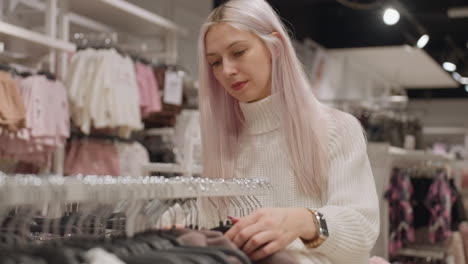 selective shopper in white knitted sweater browses neutral garments on boutique rack under warm lighting, returns polo to rack and examines others with manicured hand checking fabric texture