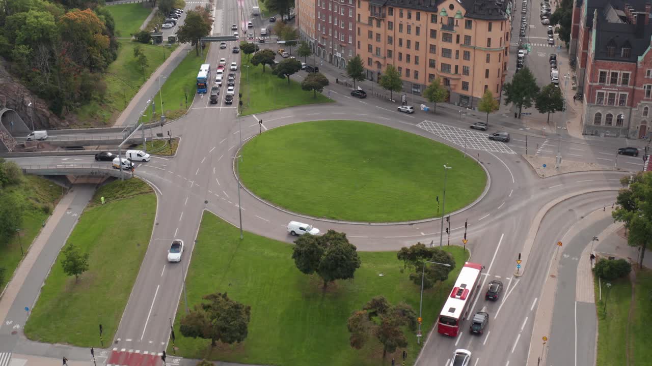 imágenes de drones de tráfico conduciendo en la rotonda de hierba verde en roslagstull, estocolmo, suecia en un día nublado con hierba verde exuberante y árboles al comienzo del otoño revelando el horizonte de la ciudad