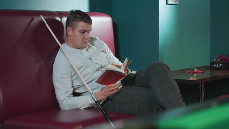 male in white shirt and plaid pants sits on red sofa in billiard room, reading book while rolling orange billiard ball in hand. cue stick leans beside him. green pool table in background