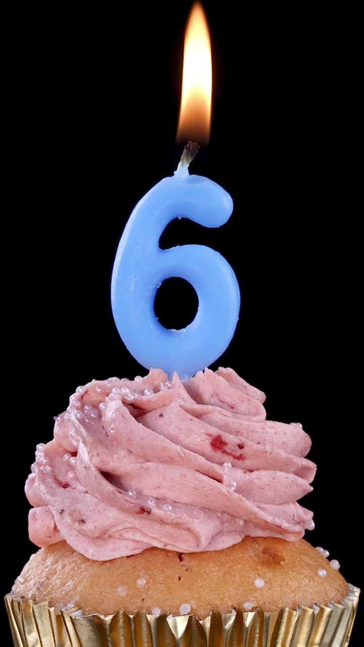 Candela blu numero sei che brucia su un cupcake alla fragola con pirottino dorato, isolata su sfondo nero.