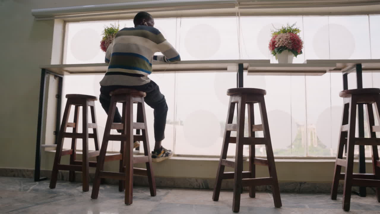 Man waiting at cafe window seat, solo diner watches street through large glass, wooden stools lined along counter with small flower vase, soft morning light creates warm silhouette, casual sweater