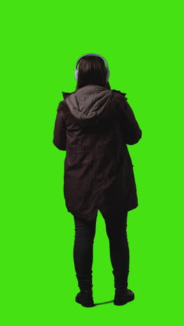 vertical video rear view full length shot of woman wearing wireless headphones streaming music from mobile phone against green screen