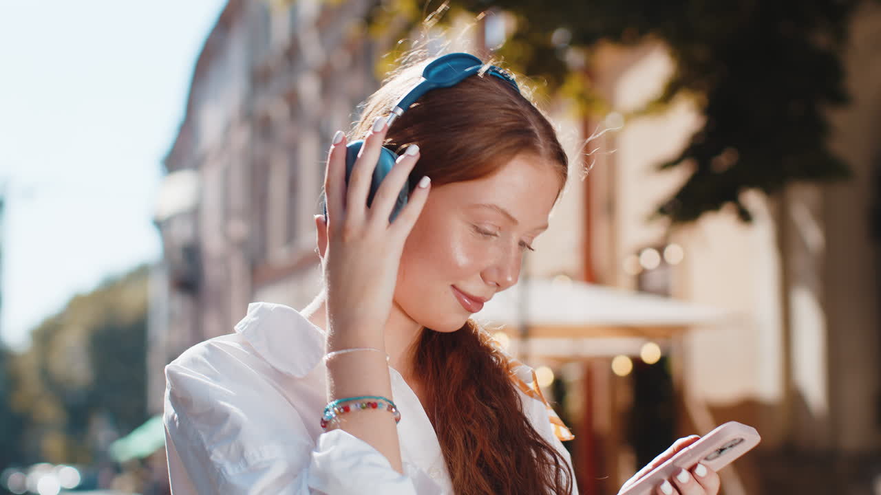 Happy teenager girl in wireless headphones choosing listening music dancing outdoors city street