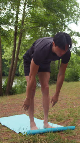 Vertical-Video-Of-Man-Wearing-Sports-Clothing-Doing-Yoga-Stretches-On-Mat-In-Forest-By-Lake-Or-River-Enjoying-Peace-And-Beauty-Of-Nature-Shot-In-Real-Time-3