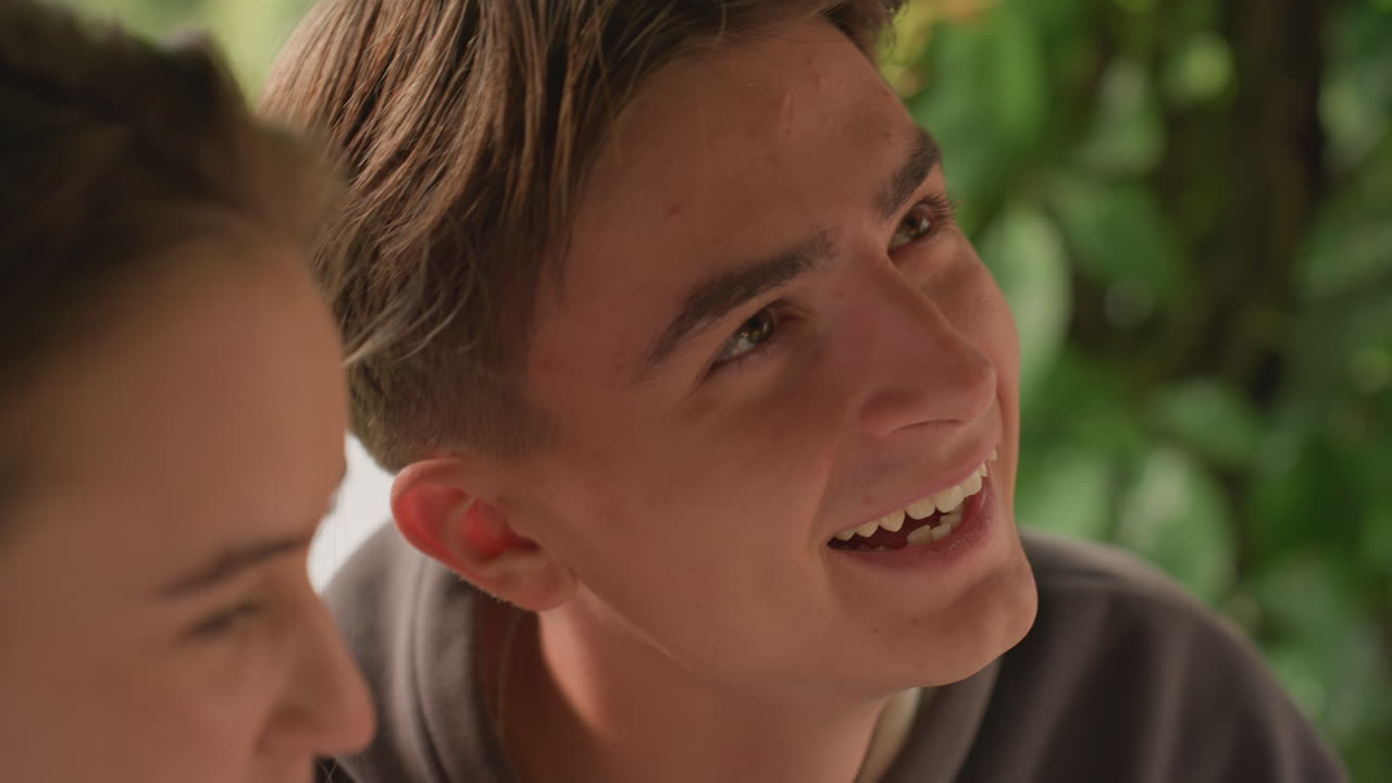 closeup caucasian teens smiling and laughing under green canopy, friends teasing each other, shy flirtation, shared joke and banter, close companionship, candid warmth and sparkling eye contact