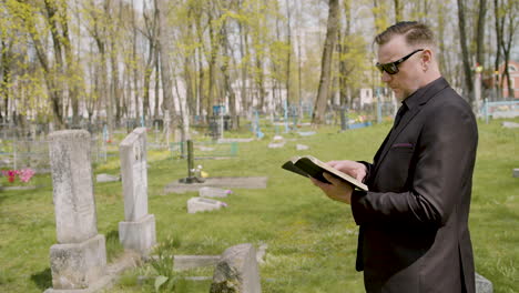 man in black suit and sunglasses reading a bible in front of a tombstone in a graveyard