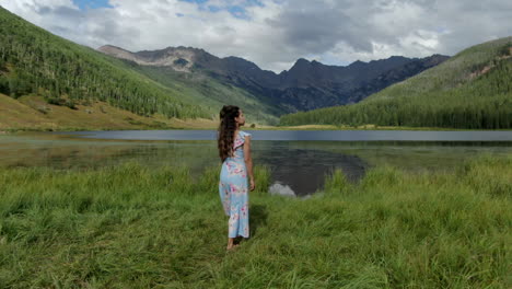 Filmische-Luftdrohne,-Die-Einer-Weiblichen-Frau,-Einem-Modell,-Einer-Schauspielerin,-Einem-Süßen-Kleid-Folgt,-Das-Mit-Breiten-Armen-Auf-Die-Piney-Lake-Ranch,-Den-Vail-Beaver-Creek,-Den-Colorado-Gore-Range,-Die-Berglandschaft,-Den-Sommernachmittag,-Regenwolken-Zugeht