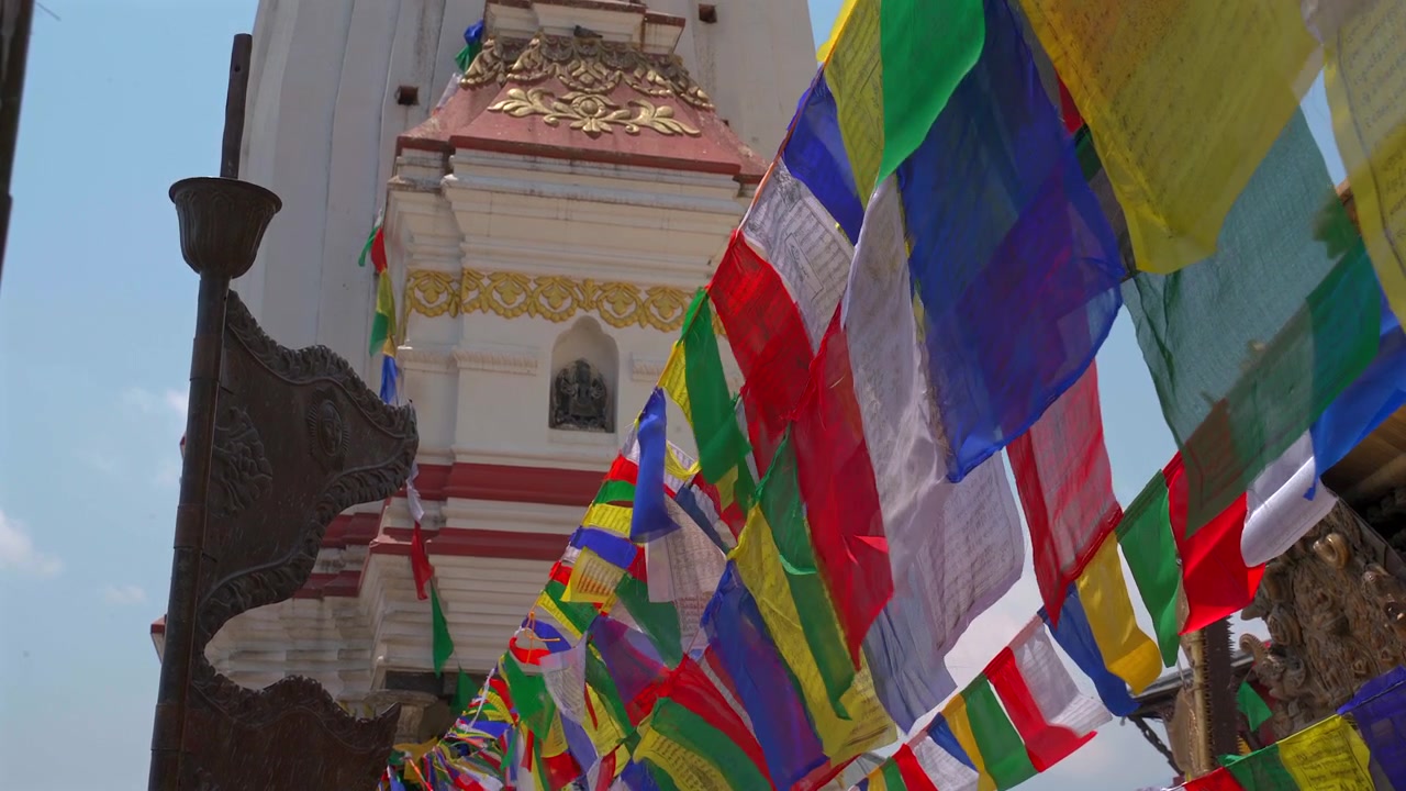 A static low-angle medium shot focusing on colorful Tibetan prayer flags fluttering heavily in the wind with a traditional white and gold temple structure in Kathmandu, Nepal.