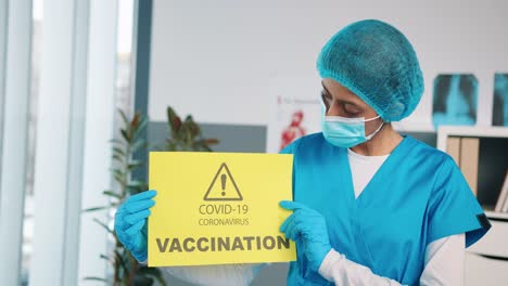 Close-Up-Portrait-Of-Hindu-Young-Woman-Specialist-Nurse-In-Protective-Gloves-And-Medical-Mask-Holding-In-Hands-Yellow-Paper-Card-Showing-Announcement-About-Covid-Vaccination,-Coronavirus-Vaccine