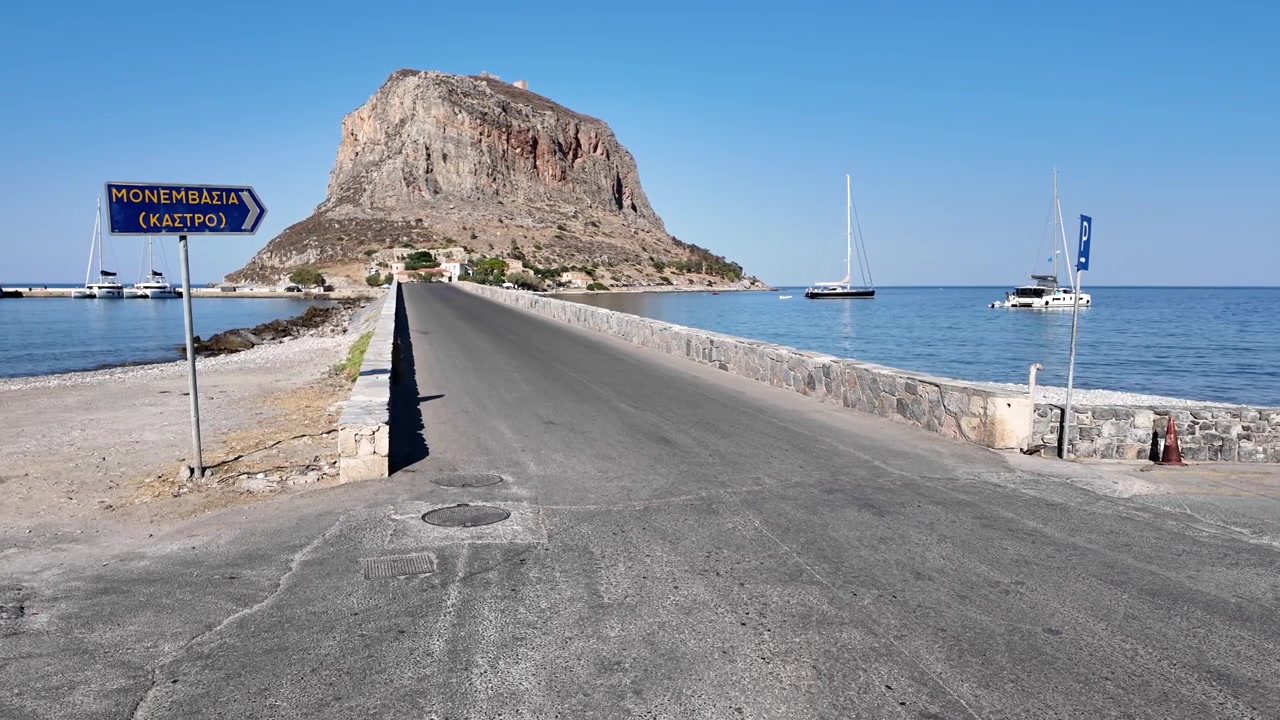 Peloponneso, Monemvasia. La strada del ponte che porta all'antica cittadella, circondata dal mare su entrambi i lati. L'imponente formazione rocciosa della cittadella spicca in una giornata di sole. Barche all'ancora.