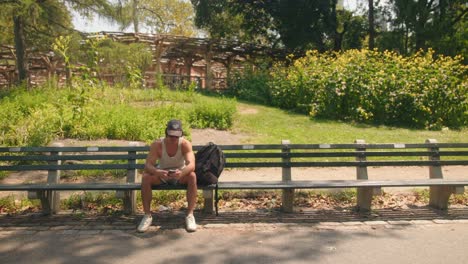 Un-Hombre-Sentado-En-Un-Banco-Del-Parque-Revisa-El-Teléfono,-Mira-Hacia-Arriba,-Parado-Y-Sale-Del-Marco-Vacío.