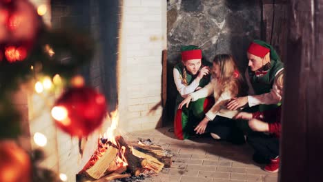 happy children and santa claus elves warm their hands near the fireplace in the room with the christmas tree