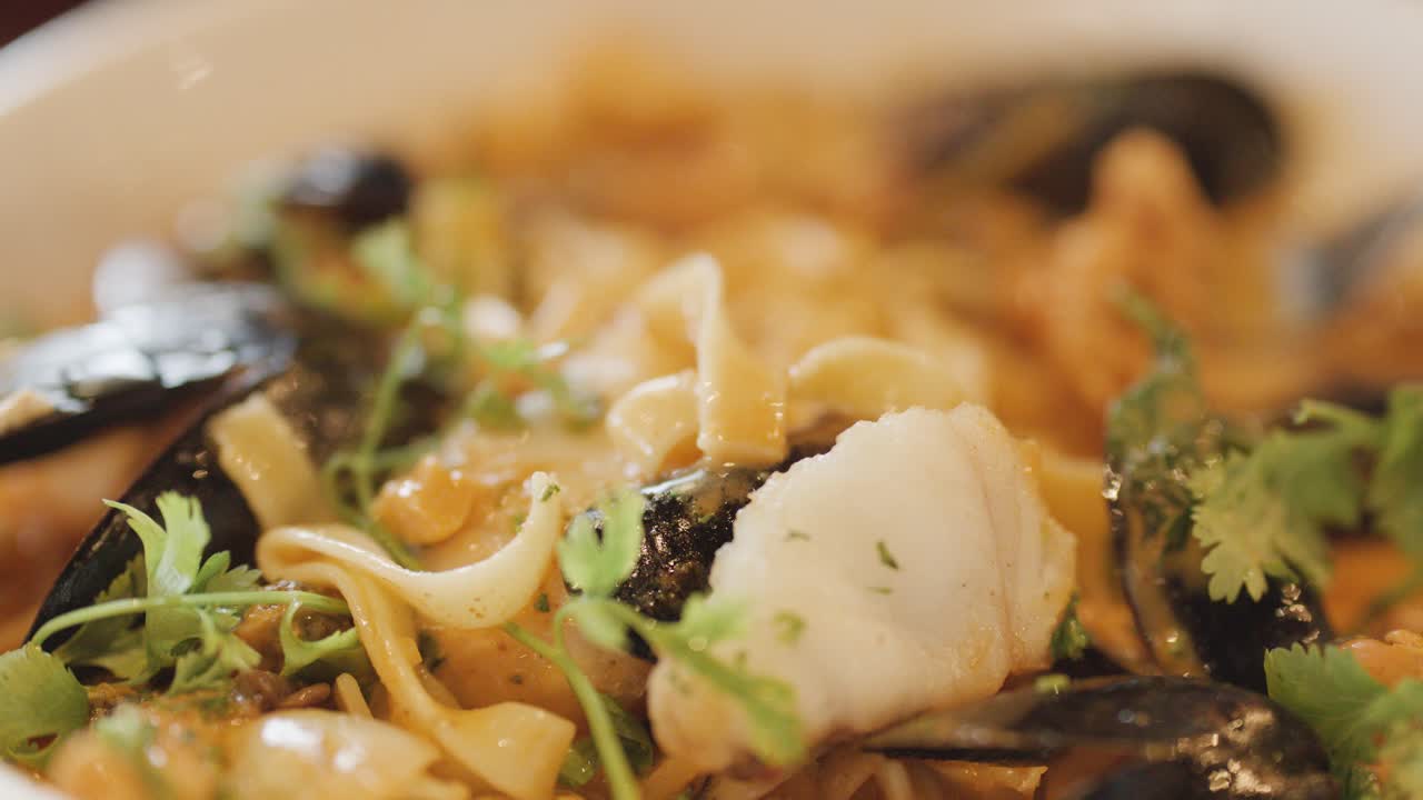Macro view of assorted seafood pasta with mussels, scallops, and fresh herbs in warm, soft lighting. Subtle camera movement highlights textures and steam