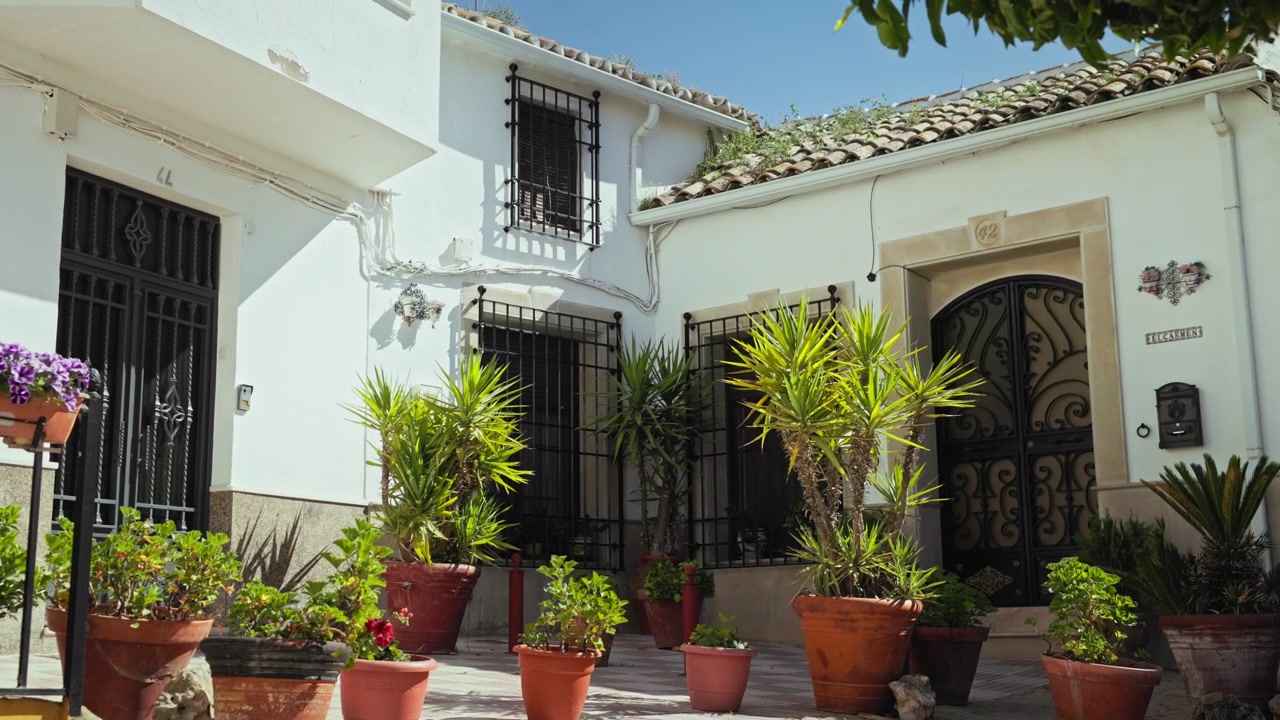 La telecamera inquadra dal basso verso l'alto piante in vaso e la facciata bianca di un cortile andaluso