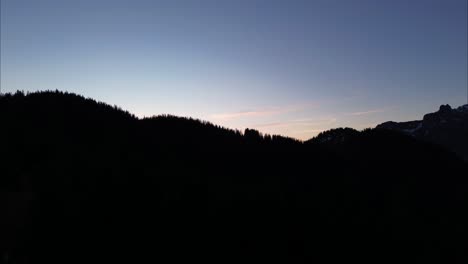 Drone-slowly-fly-over-Pine-Forest-and-Reveals-Sunrise-behind-Mountain-Range-with-Snow,-Bludenz,-Austria,-Europe