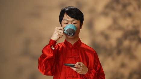 close-up view of cheerful young asian man in red traditional costume drinking tea from a ceramic cup and looking at camera