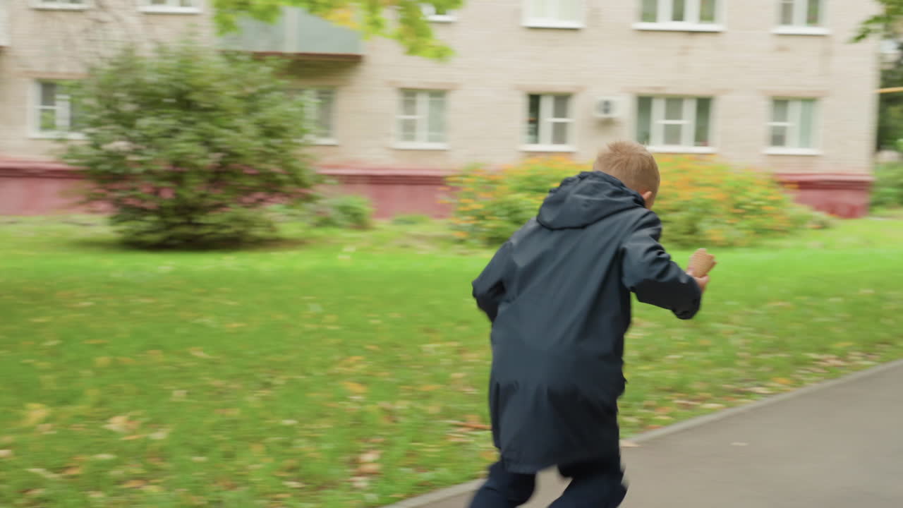 Young boy preparing for school, walking along leafy park path with backpack on bench, eating snack, wearing dark jacket, focused expression, slow deliberate pace, weekday autumn morning