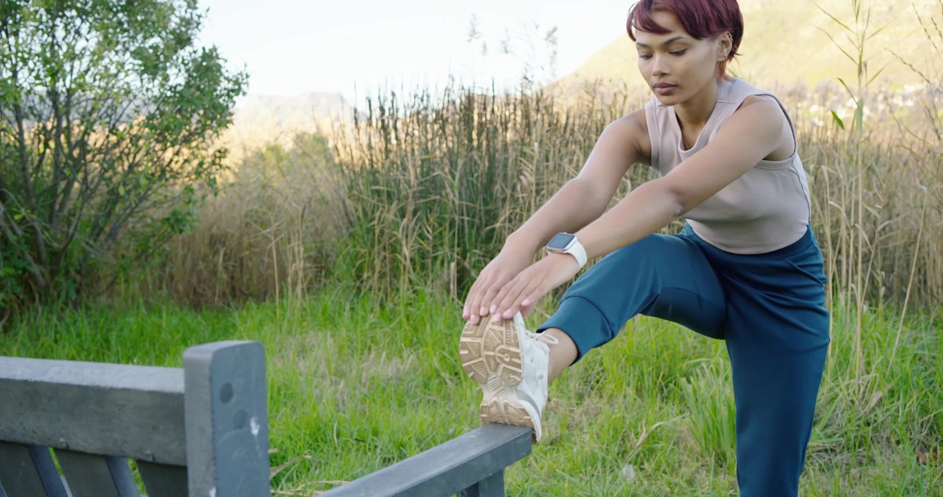 African American woman walking to bench in meadow placing foot on rail stretching for warmup