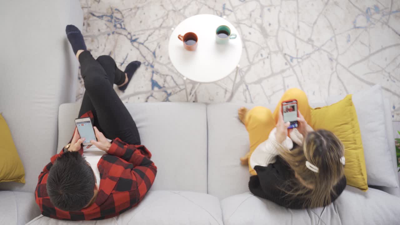 젊은 부부가 집에서 소파에 앉아서 휴대폰을 쳐다보고 있습니다.