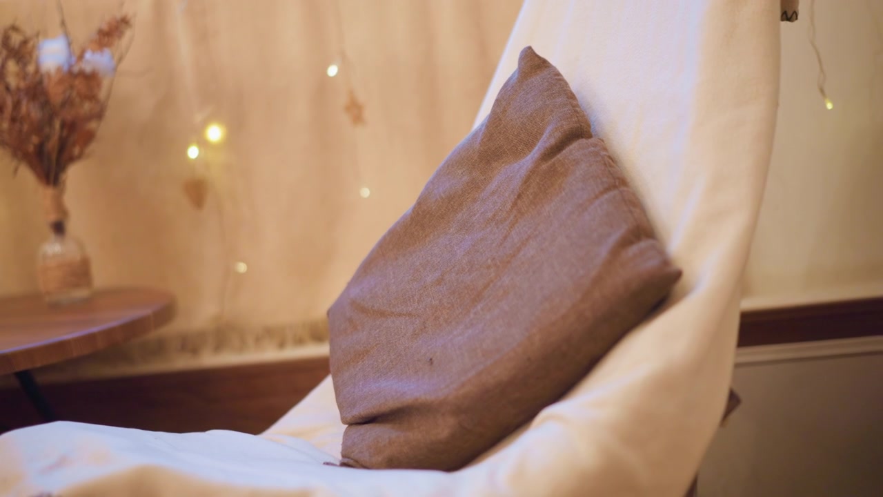 Primo piano di un cuscino in tessuto marrone appoggiato su una sedia bianco sporco con illuminazione soffusa e sfondo sfocato. La scena presenta una luce ambientale calda, lucine decorative in lontananza e un tavolo in legno.