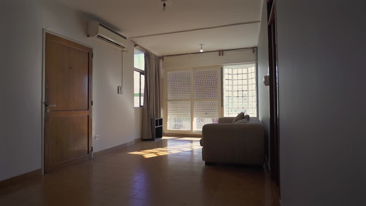 Slow camera pan reveals a clean, sunlit apartment living room with beige couch, shuttered windows, wooden door and air conditioner, minimal decor and soft natural light, slow motion.