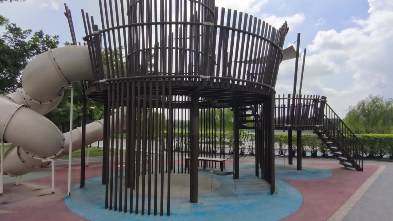 Un parque infantil vacío presenta una moderna estructura de toboganes integrada. La luminosa zona de recreo se encuentra bajo el sol del mediodía. No se ven niños jugando en este momento, creando una atmósfera tranquila.