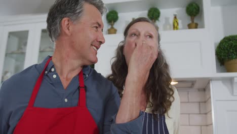 Happy-senior-caucasian-couple-in-kitchen-preparing-meal,-husband-giving-wife-a-taste-and-smiling