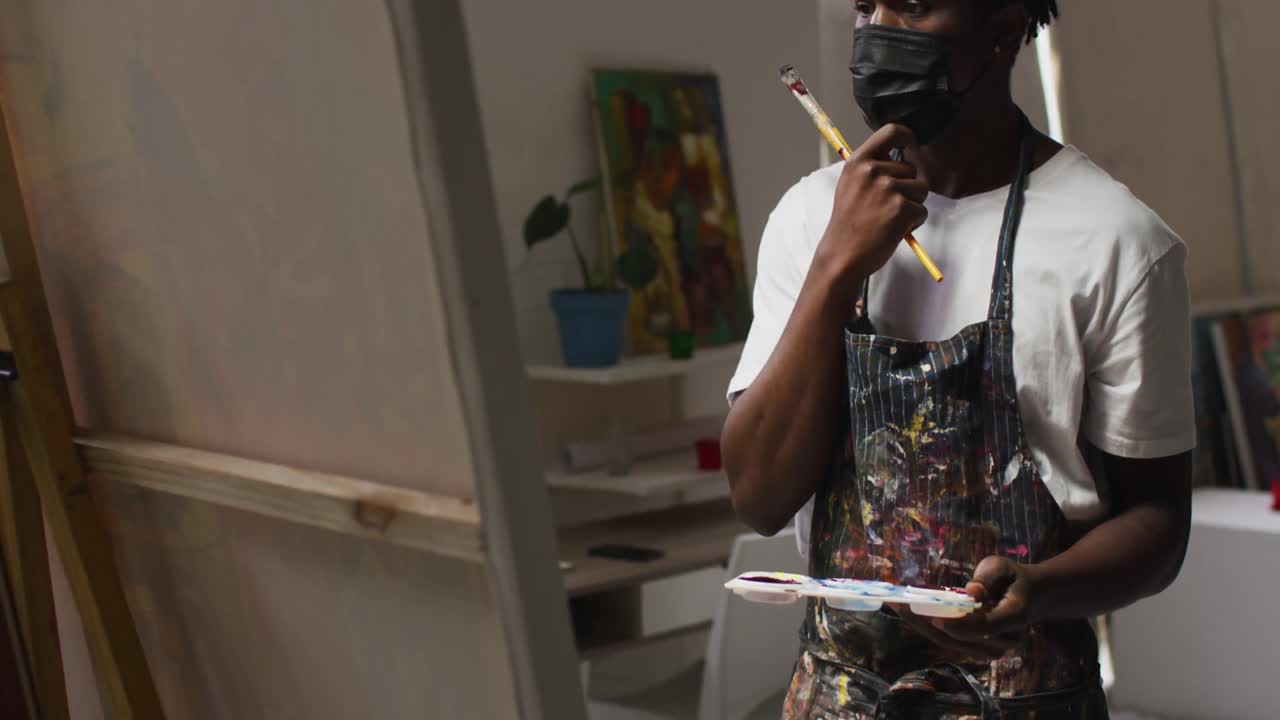 artista masculino afroamericano pensativo con pinceles y paleta en un estudio de arte