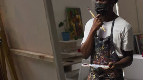 thoughtful african american male artist holding paint brushes and palette at art studio