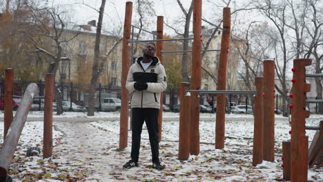 young man wearing winter jacket, standing outdoors in front of workout equipment with hands joined, performing neck twist in cold winter weather with snow and bare trees in background