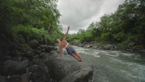 blond female on rocky bank of tropical jungle doing side plank, vasisthasana