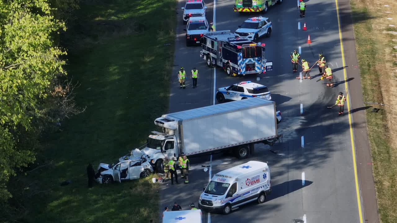 Firefighter cleaning asphalt of highway after car collision with truck. Sunny day with ambulance, police and fire department. Aerial zoom in shot. Pennsylvania, USA in autumn.