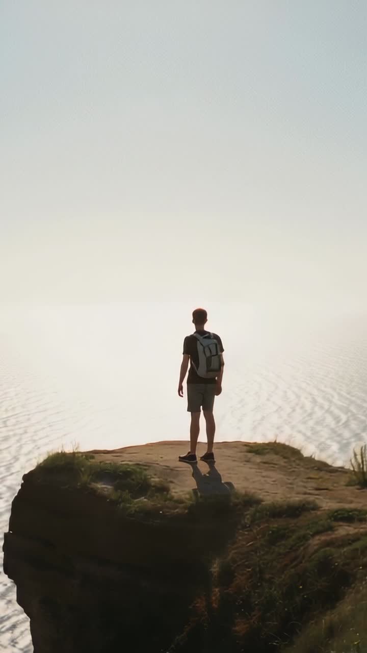 Video verticale: Raggiungendo la cima della scogliera, un escursionista si ferma, ammirando il mare in pantaloncini e con uno zaino.